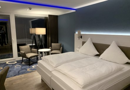 Modernes Hotelzimmer im Seehotel am Stausee, © Seehotel am Stausee Modern eingerichtetes Hotelzimmer im Seehotel am Stausee mit einem großen weiß bezogenen Doppelbett, LED-Licht, Teppich und einem Beistelltisch mit Stühlen.