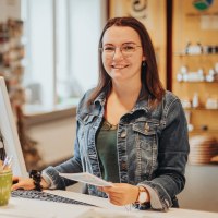 Leonie Post, Mitarbeiterin der Touristik GmbH Gerolsteiner Land, stellt sich vor.
, © Laura Kirwel Eine Frau steht an einem hohen Schreibtisch mit Computer und Tastatur vor sich und lächelt in die Kamera. Hinter ihr ist der Souveniershop und einige Prospektregale der Tourist-Information.