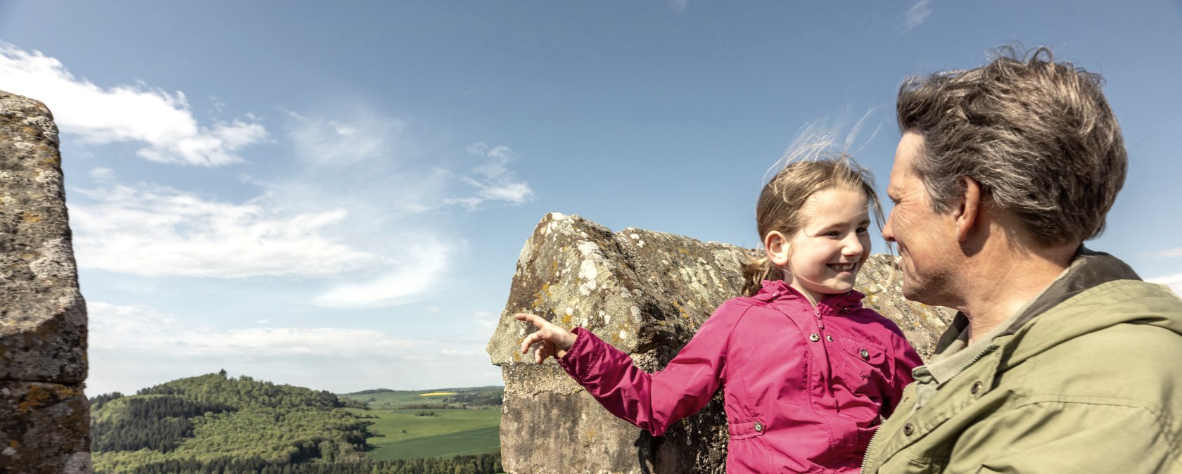 Adler & Wolfspark Kasselburg, Pelm, © Eifel Tourismus GmbH, Dominik Ketz Ein Vater trägt seine Tochter auf dem Arm auf einer Trumspitze einer Burg. Das Mädchen zeigt freudig in die Ferne.