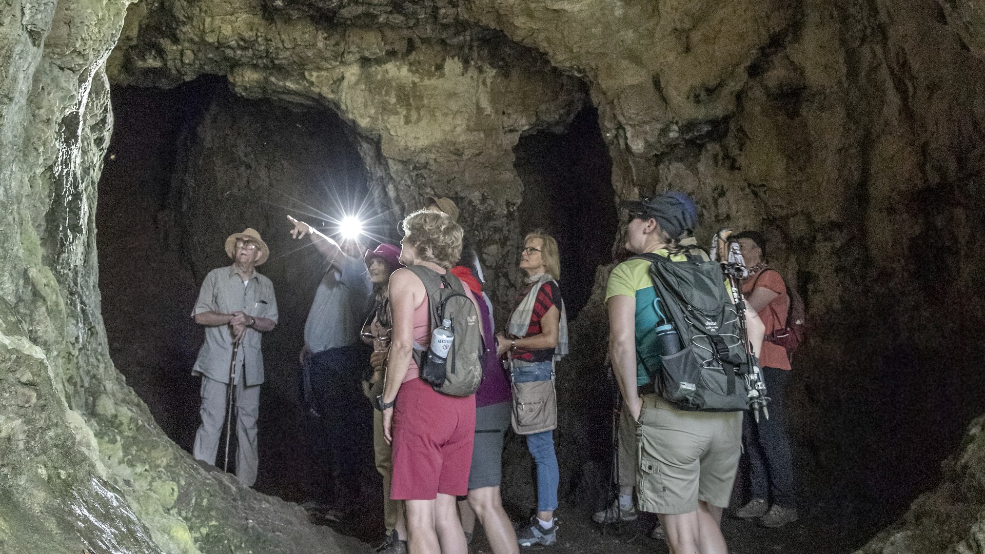 The Gerolstein Beech Hole Cave
, © Jochen Hank A hiking group of around 10 people stands in the interior of a large cave. The people are marvelling at the massive stone walls around them.