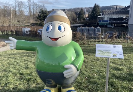 Die Willi Basalt Figur begrüßt Familien im Kurpark Stadtkyll, © Touristik GmbH Gerolsteiner Land Eine Lebensgroße Figur des Maskottchens Willi Basalt mit grünem Oberteil, grauer Hose, gelben Schuhen und einer Mütze auf dem Kopf. Die Figur lacht freundlich und zeigt in einen Park hinein.