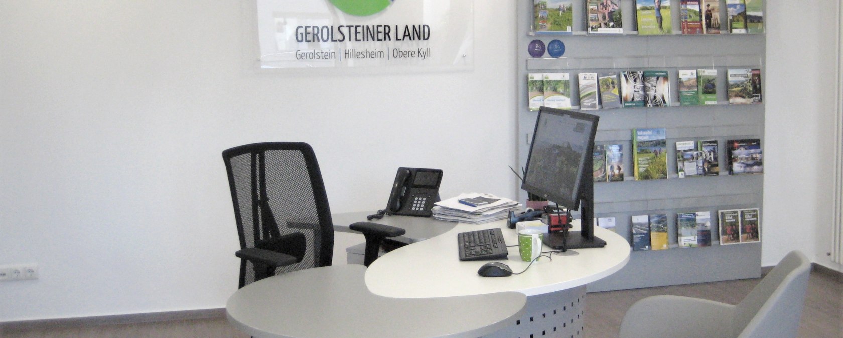 Interior of the Stadtkyll Tourist Information Centre.
, © Touristik GmbH Gerolsteiner Land Interior of the Stadtkyll Tourist Information Centre with workstation including computer, telephone, keyboard, etc., next to it a shelf unit with brochures.