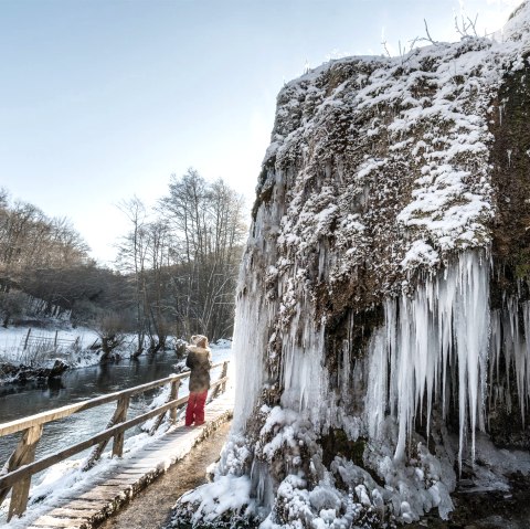 Frozen Nohner waterfall with icicles.
, © Eifel Tourismus GmbH, Dominik Ketz Frozen Nohner waterfall with icicles, a person on a footbridge, winter landscape with snow and bare trees.