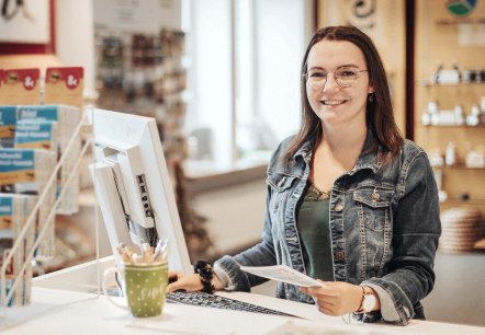 Leonie Post, employee of Touristik GmbH Gerolsteiner Land, introduces herself., © Laura Kirwel A woman stands at a high desk with a computer and keyboard in front of her and smiles at the camera. Behind her is the souvenir shop and some brochure racks from the tourist information office.