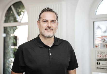 Kevin Hopp, Mitarbeiter der Touristik GmbH Gerolsteiner Land, stellt sich vor.
, © Laura Kirwel Ein Mann steht hinter einem Tresen und lächelt in die Kamera. Hinter ihr einige Fenster und Regale mit regionalen Produkten.