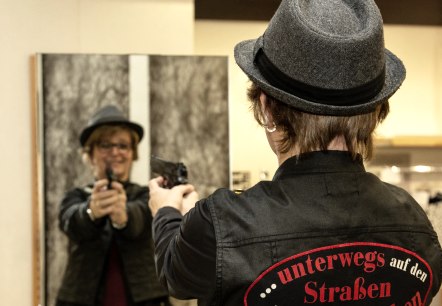 Misdaadkoningin "Dane Spur" in actie
, © Laura Kirwel Een vrouw staat voor een spiegel en richt een pistool op haar eigen spiegelbeeld. Ze draagt een grijze hoed en een jas met de inscriptie “...on the streets of crime”.