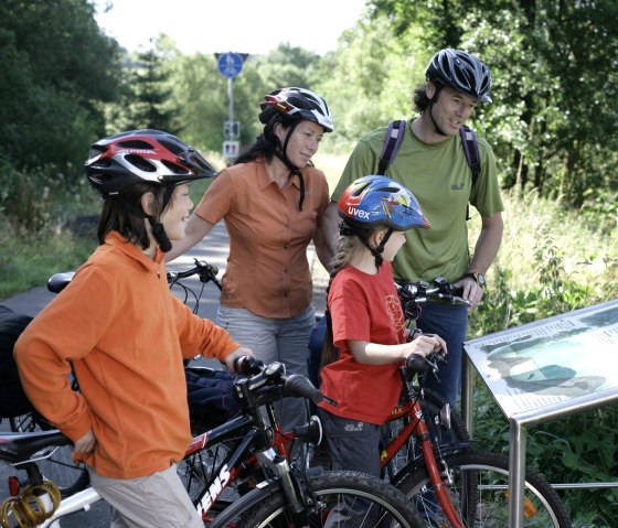 Familie mit Fahrrädern liest eine Infotafel über Biber auf dem Eifel-Ardennen-Radweg. Sie tragen Helme und sind von grüner Natur umgeben., © Eifel Tourismus GmbH/intention Familie mit Fahrrädern liest eine Infotafel über Biber auf dem Eifel-Ardennen-Radweg. Sie tragen Helme und sind von grüner Natur umgeben., © Eifel Tourismus GmbH/intention