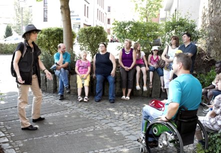 Krimispaziergang durch die Krimihauptstadt Hillesheim, © Molter Eine Gruppe voon etwa 11 Personen bestehend aus Erwachsenen und Kindern sitzt nebeneinander in einem Kreis auf einer Mauer neben einer Straße. Vor der Gruppe steht eine Gruppenführerin und erklärt etwas.