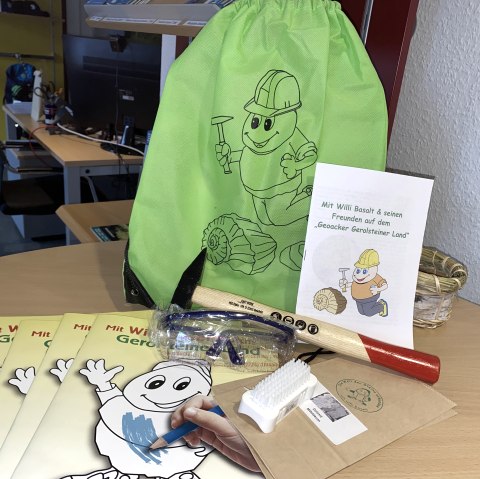 Der Forscherrucksack mit allen Werkzeugen für den Geoacker in Gerolstein., © Touristik GmbH Gerolsteiner Land Auf einem runden Holztisch liegt die Forscherausrüstung für den Gerolsteiner Geoacker: ein grüner Stoffbeutel, mehrere Malhefte, ein Hammer, eine Schutzbrille, eine Bürste und ein Infoheft.