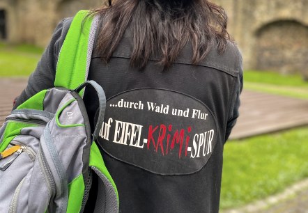 Krimi-Dame vor der Hillesheimer Stadtmauer, © Touristik GmbH Gerolsteiner Land, Gianna Kern Blick auf den Rücken einer Frau, die einen Hut und eine bedruckte Jacke sowie einen grünen Rucksack über einer Schulter trägt. Vor ihr liegt die Hillesheimer Stadtmauer.
