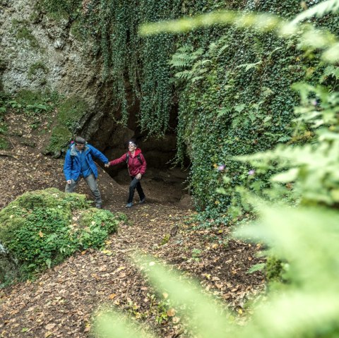 Twee wandelaars in kleurrijke jassen staan voor een grot in het met planten overwoekerde bos. De grond is bedekt met bladeren., © Eifel Tourismus GmbH, Dominik Ketz Twee wandelaars in kleurrijke jassen staan voor een grot in het met planten overwoekerde bos. De grond is bedekt met bladeren., © Eifel Tourismus GmbH, Dominik Ketz