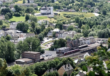 Uitzicht van bovenaf op het station van de stad Gerolstein.
, © Thomas Hendele (alle Rechte vorbehalten) Uitzicht van bovenaf op delen van de stad Gerolstein met het station, waar net een trein stopt. Rondom bomen, parkeerplaatsen en talrijke gebouwen.