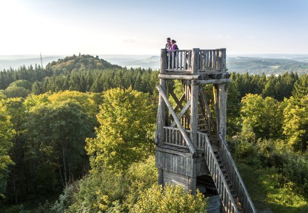 The lookout tower on the Dietzenley volcano, © Eifel Tourismus GmbH, Dominik Ketz Two hikers stand at the top of a multi-storey wooden observation tower, which juts out of a dense forest, and look into the distance.