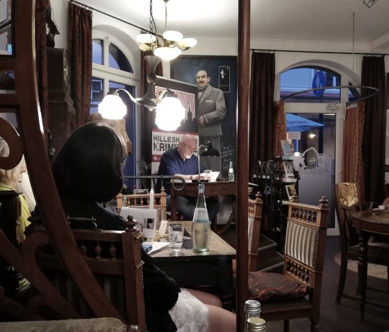 Krimilesung im Café Sherlock in Hillesheim
, © Touristik GmbH Gerolsteiner Land, Nadja Blum Schwach beleuchtes Café, in dem einige Personen sitzen und zum Autor schauen, der vorliest.