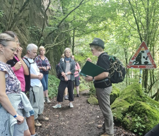 Wandergruppe auf dem Gerolsteiner Krimisteig mit krimi-Dame "Dane Spur", © Touristik GmbH Gerolsteiner Land Mehrere Wanderer stehen nebeneinander auf einem Waldpfad und schauen zu einer Dame, die Ihnen etwas erklärt oder vorliest.