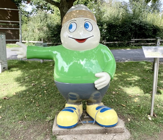 Colorful figure of mascot "Willi Basalt" with green top, blue trousers and yellow shoes stands on a meadow under a tree., © Touristik GmbH Gerolsteiner Land Colorful figure of mascot "Willi Basalt" with green top, blue trousers and yellow shoes stands on a meadow under a tree., © Touristik GmbH Gerolsteiner Land