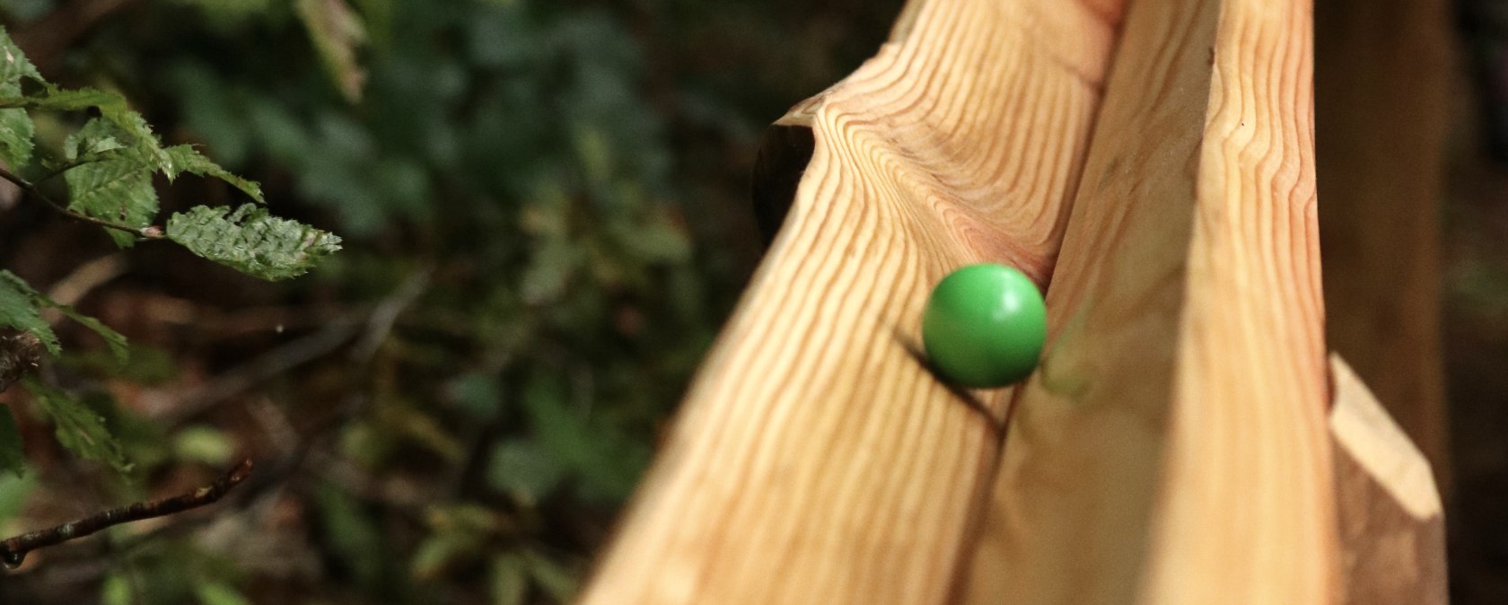 Holzkugelbahn, auf der eine grüne Kugel rollt., © Thomas Langens Zwei Holzbalken aneinander bilden ene Rille, auf der eine bunte Holzkugel rollt. Dicht daneben Laubbäume und ein Kind im Hintergrund.