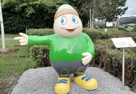Familienfreundliches Maskottchen Willi Basalt begrüßt Familien vor dem Adler- und Wolfspark Kasselburg in Pelm, © Touristik GmbH Gerolsteiner Land Statue von Maskottchen Willi Basalt mit grünem Shirt, grauer Hose und gelben Schuhen zeigt nach links. Hinter ihm Bäume und ein Parkplatz.