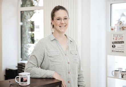 Malin Blomenkamp, Mitarbeiterin der Touristik GmbH Gerolsteiner Land, stellt sich vor.
, © Laura Kirwel Eine Frau steht hinter einem Tresen und lächelt in die Kamera. Hinter ihr einige Fenster und Regale mit regionalen Produkten.
