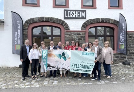 Die Presskonferenz zum Radaktionstag Grenzenlos Kyllradweg 2025 in Losheim., © Gemeinde Hellenthal Vertreter der Ortsgemeinden entlang der Strecke und Politiker stehen dicht beeinander und halten das Veranstaltungsbanner hoch.