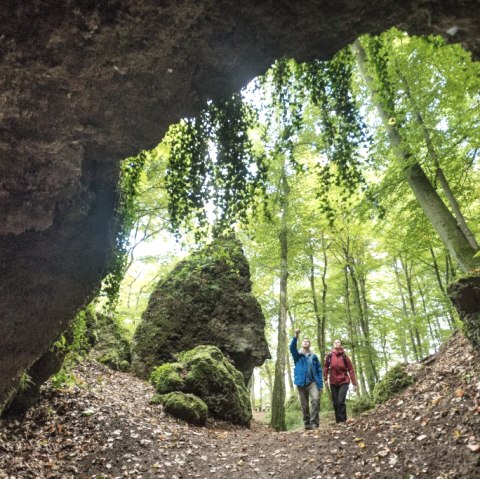 Wanderer vor den Birresborner Eishöhlen, © Eifel Tourismus GmbH, Dominik Ketz Zwei Personen wandern auf einen Höhleneingang aus Stein zu und zeigen auf die Felsen runderhum. Der Wald vor den Höhlen ist voller grün blühender hoher Bäume.