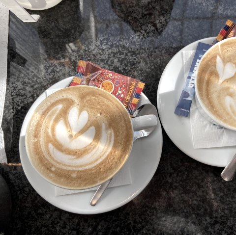 Zwei liebevoll zubereitete Tassen Cappuchino.
, © Bistro im Flecken, Julia Schneider Zwei randvoll gefüllte Tassen Cappuchino mit Milchschaumkunst auf Untertassen mit jeweils einem Löffel, Zuckerpäckchen und einem verpackten Keks.