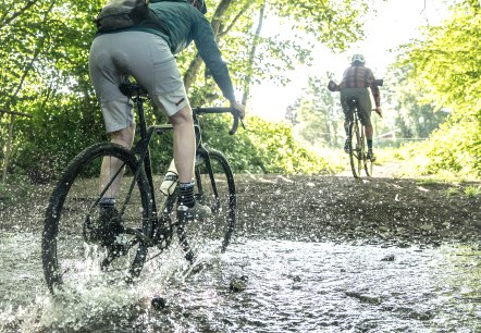 Gravelbike, © Eifel Tourismus GmbH, Dennis Stratmann Een fietser op een gravelbike rijdt door het midden van een grote fontein in een bos. Het water spat aan alle kanten op. Een andere fietser rijdt voor hem uit het bos.