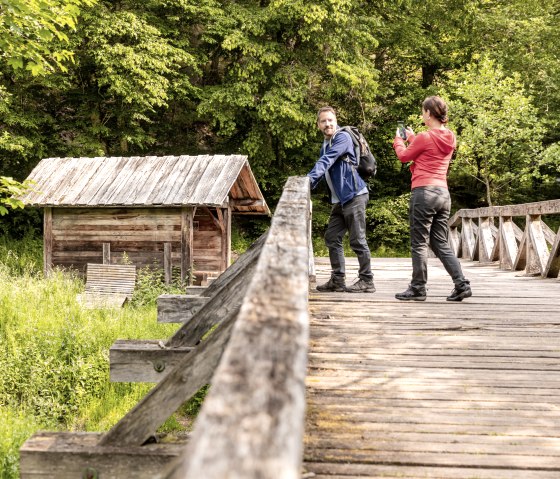 Two people are standing on a wooden bridge in front of a small wooden hut surrounded by lush greenery. One person is taking a photo., © Eifel Tourismus/AR-shapefruit AG Two people are standing on a wooden bridge in front of a small wooden hut surrounded by lush greenery. One person is taking a photo., © Eifel Tourismus/AR-shapefruit AG