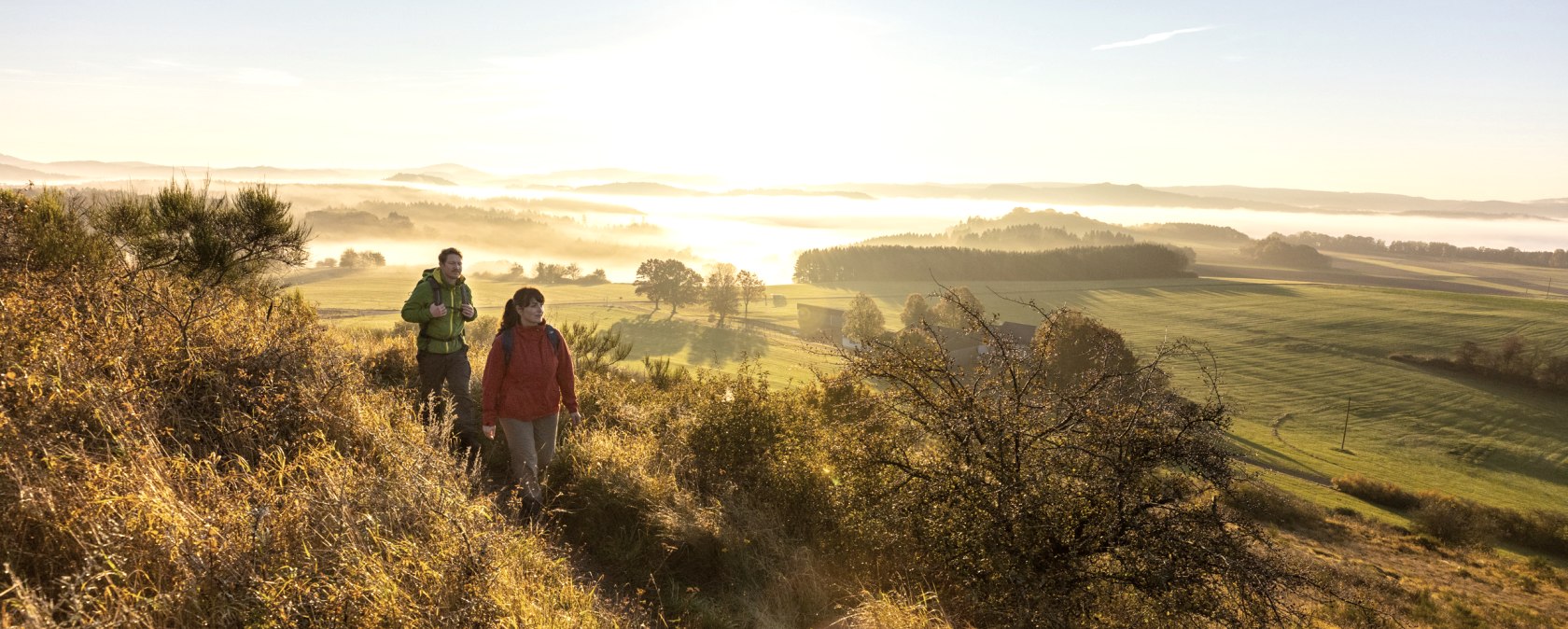Wandern in den Sonnenaufgang am Eifelsteig, Rother Kopf, © Eifel Tourismus GmbH, Dominik Ketz Zwei Wanderer auf einem Pfad in einer hügeligen Landschaft bei Sonnenaufgang.