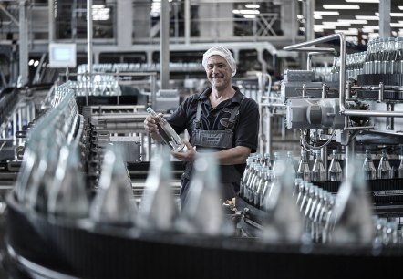 Arbeiter in der produktionsabteilung des Gerolsteiner Brunnens.
, © Gerolsteiner Brunnen GmbH Ein Arbeiter in einer Abfüllanlage überprüft eine Wasserflasche. Er trägt Arbeitskleidung und eine Haube. Um ihn herum sind viele Flaschen auf einem Förderband.