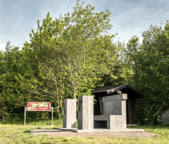 A stone monument in the shape of a throne stands next to an information board in the greenery. Trees and a small building can be seen in the background., © Eifel Tourismus GmbH, Dominik Ketz A stone monument in the shape of a throne stands next to an information board in the greenery. Trees and a small building can be seen in the background., © Eifel Tourismus GmbH, Dominik Ketz