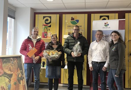 Frank Reuter, Managing Director of Touristik GmbH Gerolsteiner Land, employee Elvira Weimer and the three winners of the competition held on the opening day of the Gerolstein forest marble run.
, © Touristik GmbH Gerolsteiner Land Five people stand side by side in front of a souvenir shelf at the Gerolstein Tourist Information Office and smile for the camera. Three of them proudly hold their prizes in their hands.