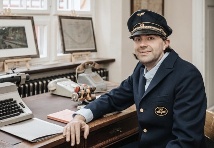 "Bahnhofsvorsteher" Andreas Kurth sitzt an seinem Schreibtisch.
, © Andreas Kurth Mann in historischer Schaffnerkleidung sitzt an einem Schreibtisch, auf dem eine Schreibmaschine, Stempel und ein antikes Telefon stehen und schaut in die Kamera.