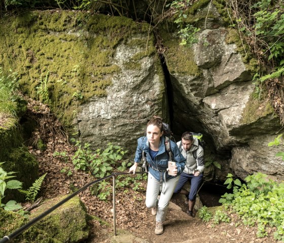 Two hikers emerge from a moss-covered cave on a forest path. They are carrying rucksacks and are surrounded by green vegetation., © Eifel Tourismus GmbH, Dominik Ketz Two hikers emerge from a moss-covered cave on a forest path. They are carrying rucksacks and are surrounded by green vegetation., © Eifel Tourismus GmbH, Dominik Ketz