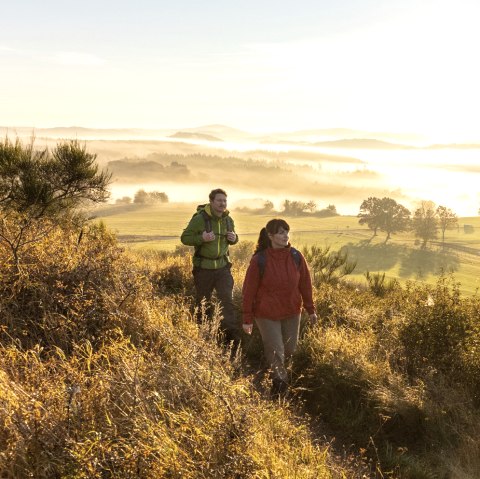 Wandern in den Sonnenaufgang am Eifelsteig, Rother Kopf, © Eifel Tourismus GmbH, Dominik Ketz Zwei Wanderer auf einem Pfad in einer hügeligen Landschaft bei Sonnenaufgang.