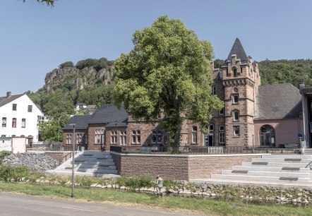 Buitengevel van het station van Gerolstein met het toeristeninformatiekantoor van Gerolstein.
, © Jochen Hank Buitengevel van het station van Gerolstein achter een dichtbegroeide boom, voor de dolomietrotsen en aan de oever van de rivier de Kyll.