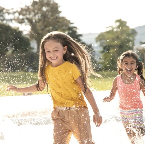 Zwei Mädchen laufen freudig durch ein Wasserbecken., © Canva Zwei Mädchen laufen lachend hintereinander durch ein Wasserbecken und das Wasser spritzt in alle Richtungen.