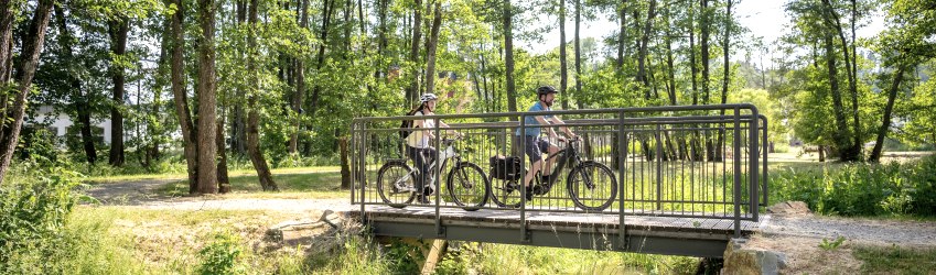 Zwei Radfahrer auf dem Kyllradweg im Kurpark Stadtkyll.
, © Eifel Tourismus GmbH, Dominik Ketz Ein Mann und eine Frau fahren auf E-Bikes über eine kleine Brücke im grünen Kurpark Stadtkyll.