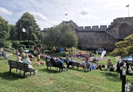 Veranstaltung im Parkgelände vor der Hillesheimer Stadtmauer
, © Touristik GmbH Gerolsteiner Land, Esther Erharter zahlreiche Personen sitzen auf Stühlen und Decken auf einer Wiese im Park und schauen zur kleinen Bühne mit Musikverein und Lesern vor der Hillesheimer Stadtmauer