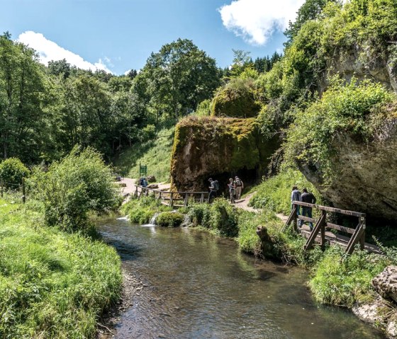 Ein Wanderweg führt entlang eines kleinen Flusses, umgeben von üppigem Grün und Felsen. Menschen spazieren auf einem Holzsteg., © Achim Meurer Ein Wanderweg führt entlang eines kleinen Flusses, umgeben von üppigem Grün und Felsen. Menschen spazieren auf einem Holzsteg., © Achim Meurer