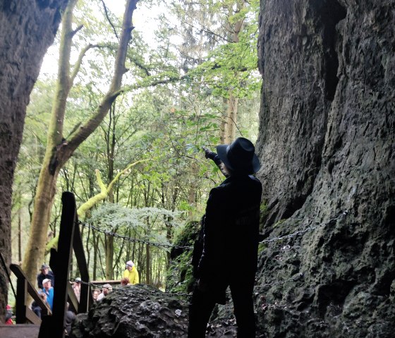Gerolsteiner Krimisteig, Buchenlochhöhle, © Dorita Molter-French Steinernder Eingang in eine Höhle, in dem eine Person steht und eine Pistole auf den Wald vor der Höhle richtet. Ein Treppe führt hinauf zu Höhle und mehrere Personen stehen am Treppenanfang.