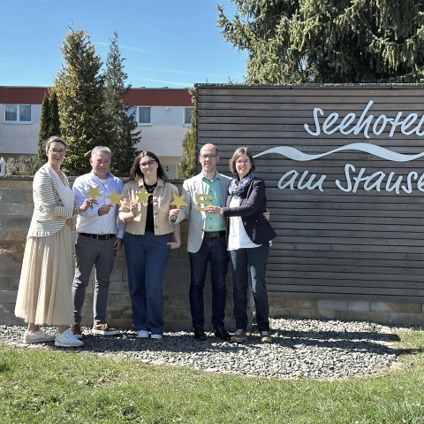 Herr Schinagl, Frau Funke und die Touristik GmbH Gerolsteiner Land freuen sich über das tolle Sterneergebnis., © Seehotel am Stausee Fünf Personen stehen vor der Fassade des Seehotels am Stausee in gerolstein und halten jeweils 4 Sterne und ein "S" für die Sterneklassifizierung hoch.