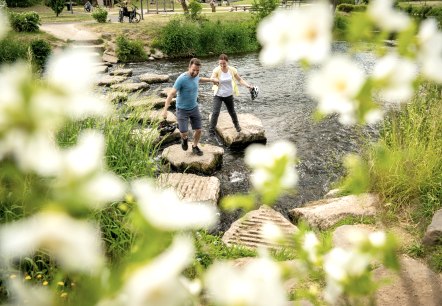 Two people cross the Kyll river in Gerolstein spa gardens using stepping stones.
, © Eifel Tourismus GmbH, Dominik Ketz A man and woman with helmets in their hands cross the Kyll river in Gerolstein spa gardens using stepping stones.