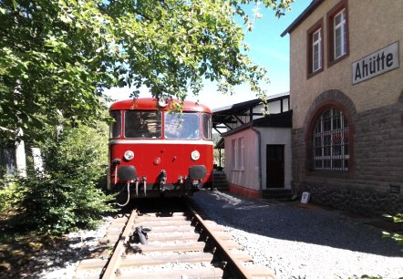 Historische rode trein voor het museumstation Ahütte.
, © Andreas Kurth Een historische rode trein staat op de sporen naast een oud stationsgebouw met het opschrift “Ahütte” op de muur.