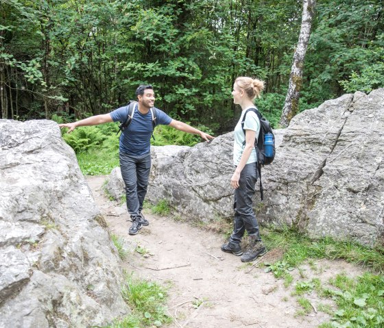 Two hikers are standing between large rocks in the forest and talking. They are carrying rucksacks and wearing hiking gear., © Eifel-Tourismus GmbH, shapefruit AG A. Röser Two hikers are standing between large rocks in the forest and talking. They are carrying rucksacks and wearing hiking gear., © Eifel-Tourismus GmbH, shapefruit AG A. Röser