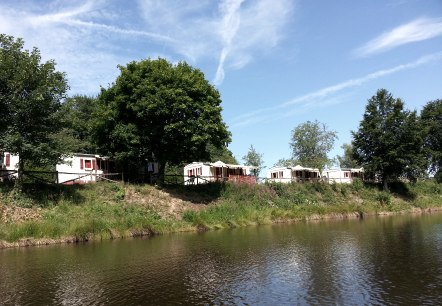 Het stuwmeer in Landalpark Wirfttal met enkele vakantiehuizen aan de oever., © Tourist Information Gerolsteiner Land Het stuwmeer in Landalpark Wirfttal met enkele kleine witte identieke vakantiehuisjes aan de oever op een weiland. Tussen wat bomen en een blauwe lucht.