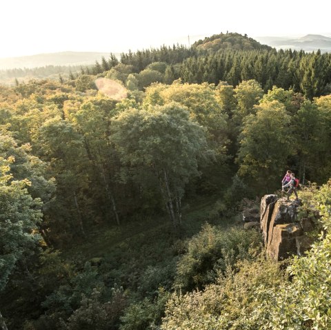 Two people are standing on a rock surrounded by a dense forest. Hills and a wide view over the landscape can be seen in the background., © Eifel Tourismus GmbH, D. Ketz Two people are standing on a rock surrounded by a dense forest. Hills and a wide view over the landscape can be seen in the background., © Eifel Tourismus GmbH, D. Ketz