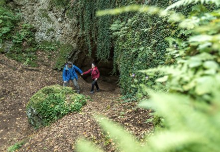 Zwei Wanderer in bunten Jacken stehen vor einer mit Pflanzen bewachsenen Höhle im Wald. Der Boden ist mit Laub bedeckt., © Eifel Tourismus GmbH, Dominik Ketz Zwei Wanderer in bunten Jacken stehen vor einer mit Pflanzen bewachsenen Höhle im Wald. Der Boden ist mit Laub bedeckt., © Eifel Tourismus GmbH, Dominik Ketz