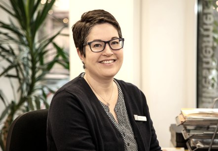 Nadja Blum, employee at Touristik GmbH Gerolsteiner Land, introduces herself.
, © Laura Kirwel A woman sits at a desk and smiles at the camera. Behind her is a large palm tree.