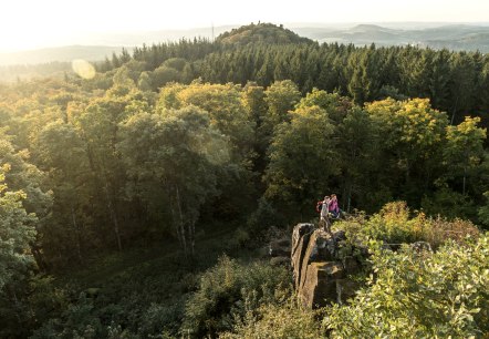 Two people are standing on a rock, surrounded by dense forest. In the background, hills and a sweeping view of the landscape can be seen.
, © Eifel Tourismus GmbH, Dominik Ketz Two people are standing on a rock overlooking a forest and hills in the background.